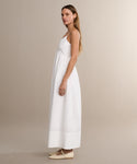 A woman in profile stands against a beige background, wearing the Peninsula Dress—a long, sleeveless white sundress made from Italian washed poplin. Her hair is straight and loose, and her expression is neutral.
