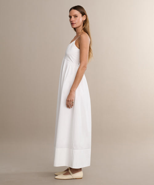 A woman in profile stands against a beige background, wearing the Peninsula Dress—a long, sleeveless white sundress made from Italian washed poplin. Her hair is straight and loose, and her expression is neutral.