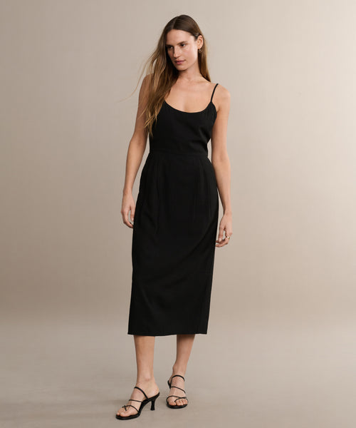 A woman poses naturally against a beige backdrop in the Rove Dress, a sleeveless black midi with thin straps, styled with strappy heels and loose hair.