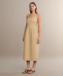 A woman with straight, light brown hair and a calm expression models the Rove Dress—a sleeveless, light beige cotton spring dress with a square neckline—paired with brown heeled sandals in a neutral setting.