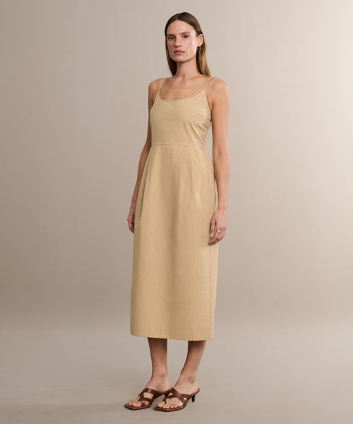 A woman with straight, light brown hair and a calm expression models the Rove Dress—a sleeveless, light beige cotton spring dress with a square neckline—paired with brown heeled sandals in a neutral setting.