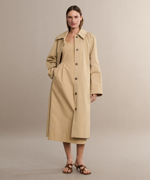 A woman stands against a plain background wearing the Rove Dress under a beige trench coat and brown sandals, with one hand in her pocket and a neutral expression.