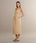 A woman with straight, shoulder-length hair wears the Rove Dress—a beige, sleeveless cotton spring dress with thin straps and pockets—in a neutral studio, paired with brown heeled sandals.