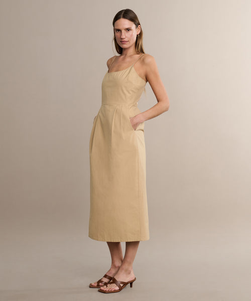 A woman with straight, shoulder-length hair wears the Rove Dress—a beige, sleeveless cotton spring dress with thin straps and pockets—in a neutral studio, paired with brown heeled sandals.