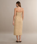 A woman with straight light brown hair faces away from the camera, wearing the Rove Dress—a khaki cotton spring dress with thin straps and a fitted waist—paired with brown heeled sandals against a neutral background.