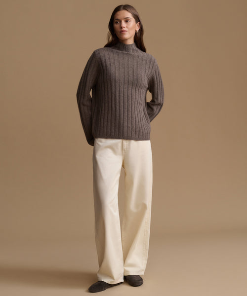 A woman stands against a warm beige studio background wearing a ribbed knit mock-neck sweater in dark taupe-brown, cream wide-leg trousers, and dark slip-on shoes.