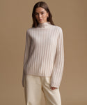 A woman modeling a cream-colored ribbed knit mock neck sweater and matching cream trousers against a solid beige studio background.