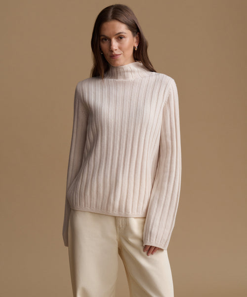 A woman modeling a cream-colored ribbed knit mock neck sweater and matching cream trousers against a solid beige studio background.