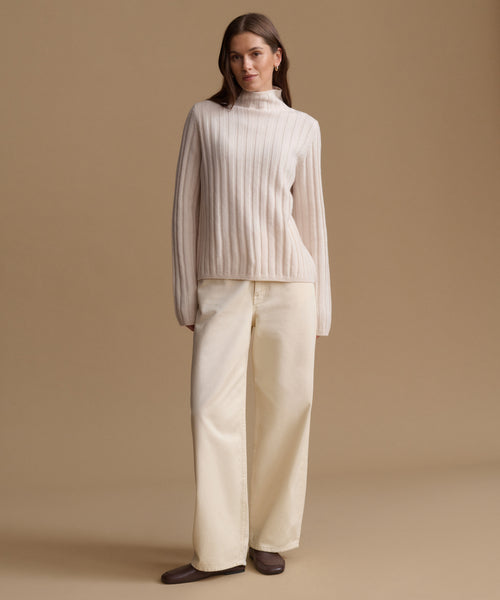 A woman stands against a warm beige studio background wearing a cream ribbed mock-neck sweater and matching cream wide-leg trousers, with dark brown shoes.