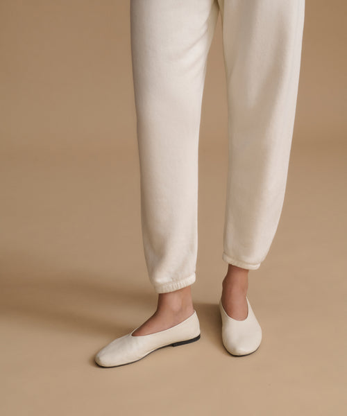 A person wearing cream-colored jogger pants and matching flat ballet flats, standing against a solid beige studio background.