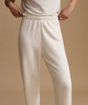 A person wearing cream-colored knit lounge pants with an elastic waistband, shown from the waist down against a warm beige studio background.