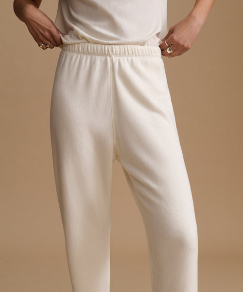 A person wearing cream-colored knit lounge pants with an elastic waistband, shown from the waist down against a warm beige studio background.