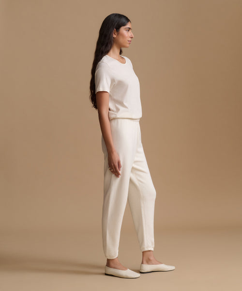 A woman in a side profile wearing a matching cream short-sleeve top and relaxed-fit cream pants, paired with cream flat shoes, against a solid beige studio background.