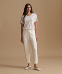 A woman modeling a matching cream-colored lounge set consisting of a short-sleeve crewneck t-shirt and relaxed-fit ankle-length pants, paired with cream flat shoes, against a solid beige studio backdrop.