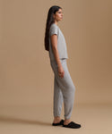 A woman stands in profile wearing a light gray short-sleeve cropped top and matching jogger-style pants, paired with black slip-on shoes, against a plain warm beige studio background.