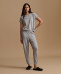 A person wearing light gray heather jogger sweatpants with an elastic waistband and ankle cuffs, paired with a matching gray top and black slip-on slippers, against a warm beige studio background.
