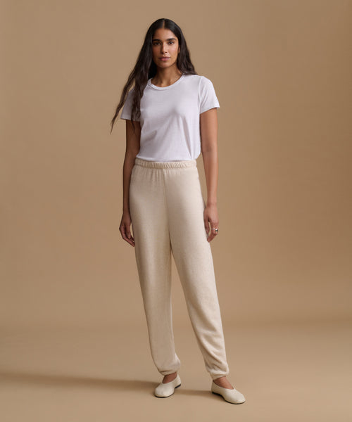 A woman stands against a warm beige studio background wearing a white short-sleeve crewneck T-shirt, cream-colored knit pants, and cream flats.