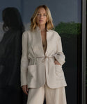 A woman with blonde hair wears the Scout Jacket—a lightweight wool piece with large pockets and a waist tie—standing before a glass wall, her reflection and greenery behind her, perfectly showcasing this wardrobe essential.