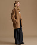 A woman stands in profile wearing a camel brown teddy coat over a white top and dark wide-leg trousers, photographed against a plain warm beige studio background.