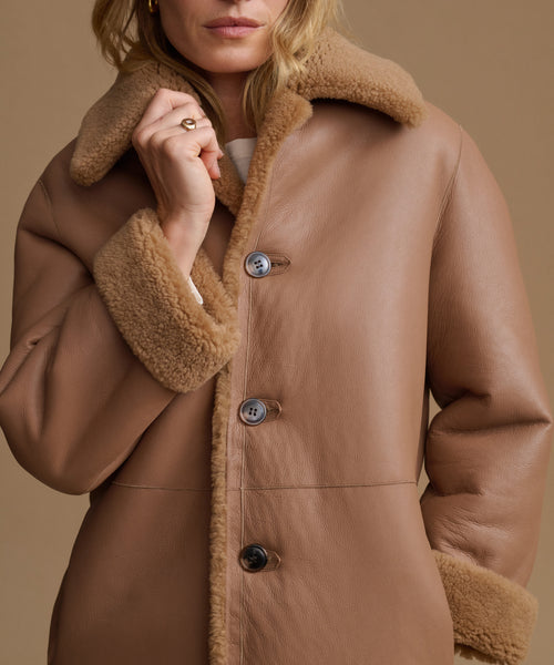 A close-up of a woman wearing a camel brown shearling-lined coat with three dark buttons, set against a solid warm beige background.