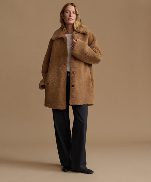 A woman stands against a warm beige studio background wearing a camel brown teddy-textured coat over a white top and dark charcoal wide-leg trousers.