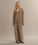 A person with long, wavy blonde hair and a neutral expression stands against a plain background wearing the Simone V-Neck sweater, wide-legged beige trousers, black sandals, and a black choker necklace.