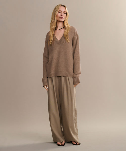 A person with long, wavy blonde hair and a neutral expression stands against a plain background wearing the Simone V-Neck sweater, wide-legged beige trousers, black sandals, and a black choker necklace.