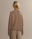 A person with blonde hair, wearing the loose-fitting Simone V-Neck brown sweater and beige pants, stands with their back to the camera against a plain beige background.