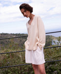 A person with short brown hair wearing the Simone V-Neck in ivory fine cotton merino and white shorts stands outdoors by a railing, surrounded by greenery and a cloudy sky—a versatile luxury layer for any season.