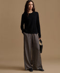A model wearing high-waisted, wide-leg trousers with vertical brown, beige, and gray stripes, paired with a black long-sleeve top and black pointed shoes, set against a solid tan studio background.