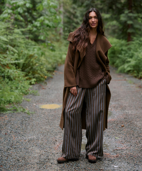 A woman stands on a gravel path in a forest wearing a brown long coat, brown knit sweater, brown-and-gray striped wide-leg pants, and brown shoes.