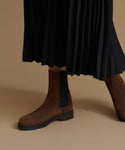 A close-up of a person wearing brown suede Chelsea boots with black elastic side panels, paired with a black pleated midi skirt, against a solid beige studio background.