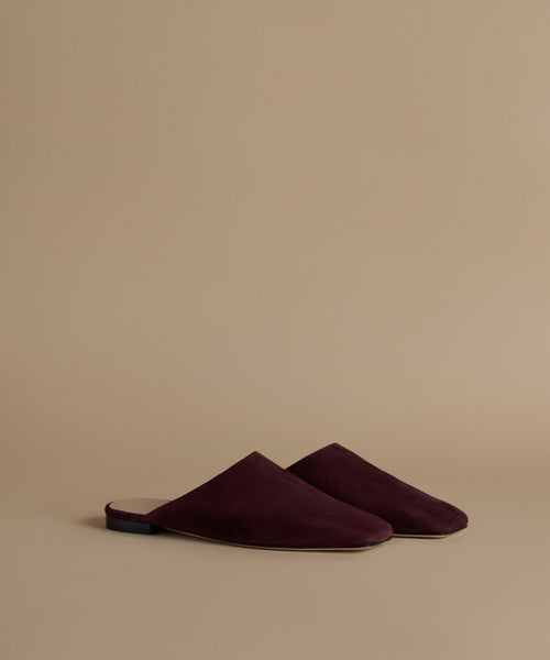 A pair of deep burgundy suede mule slippers on a solid beige background.