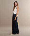 A woman with long hair stands sideways against a neutral background, wearing a white sleeveless top, black Summer Demi Pant with linen texture, cream flats, and holding a woven bag behind her back.