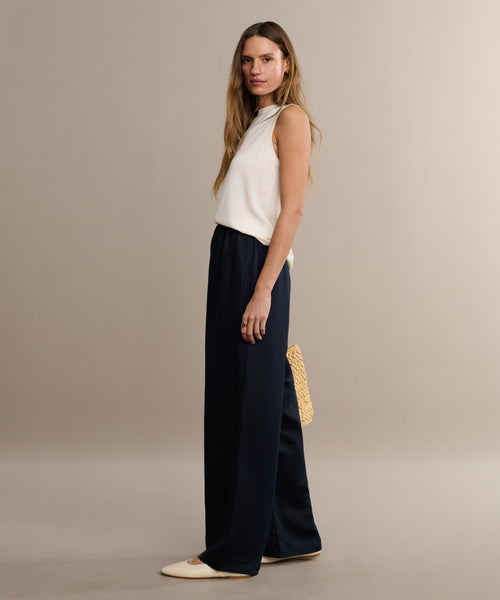 A woman with long hair stands sideways against a neutral background, wearing a white sleeveless top, black Summer Demi Pant with linen texture, cream flats, and holding a woven bag behind her back.