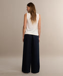 A woman with long brown hair faces away, wearing a sleeveless white top and the wide-leg black Summer Demi Pant. The pants have a subtle linen texture, and she stands against a plain neutral background.