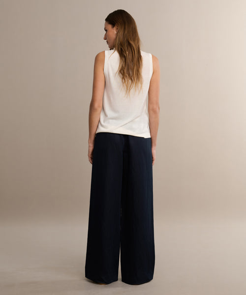 A woman with long brown hair faces away, wearing a sleeveless white top and the wide-leg black Summer Demi Pant. The pants have a subtle linen texture, and she stands against a plain neutral background.