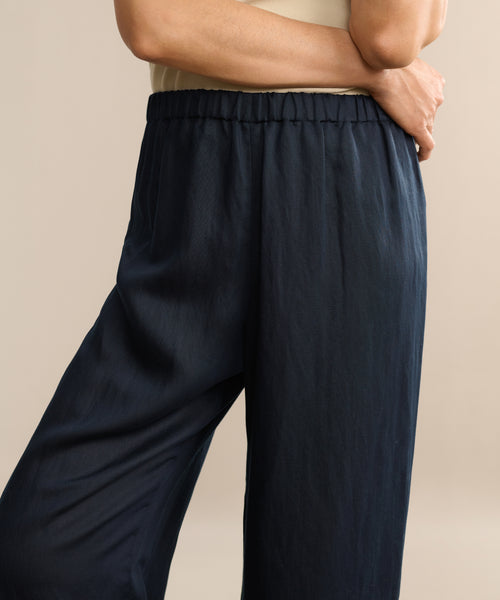 A person stands against a neutral background with one arm bent, wearing a sleeveless beige top and the Summer Demi Pant, which features a loose fit and linen texture.