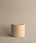 The Woven Storage Basket, made from banana leaf and featuring two handles, rests on a light brown surface with a matching background—ideal for adding style to your homes storage solutions.