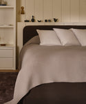 A modern, minimal bedroom features a neatly made bed with three white Adler Pillows and a light beige blanket, set against built-in shelves displaying decor and books.