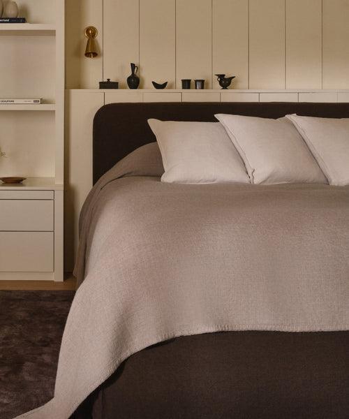 A modern, minimal bedroom features a neatly made bed with three white Adler Pillows and a light beige blanket, set against built-in shelves displaying decor and books.