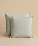 Two Adler Pillows in light gray linen blend rest against a beige background, with one slightly overlapping the other, capturing timeless Belgian-inspired style.
