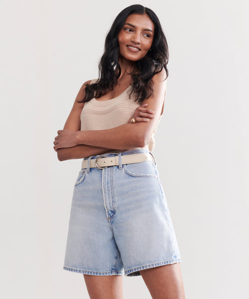 A woman with long dark hair wears a beige tank top and the Stella Short by AGOLDE, paired with a white belt. She stands with arms crossed, smiling and looking slightly to the side against a plain light background.