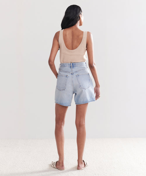 A woman stands on a light-colored carpet, facing away from the camera, wearing vintage high-waisted Stella Shorts, a beige sleeveless low-back top, and flat open-toe sandals.