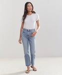 A woman stands against a plain background, wearing a white t-shirt, black strappy sandals, and the High Rise Stovepipe Jean. She looks slightly to the side with one hand in her pocket.