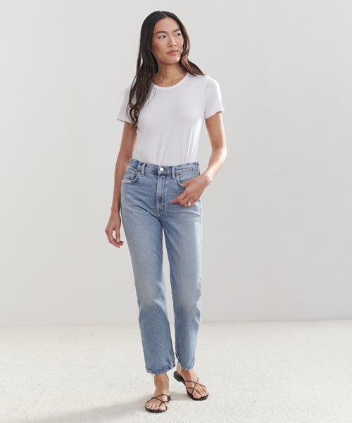 A woman stands against a plain background, wearing a white t-shirt, black strappy sandals, and the High Rise Stovepipe Jean. She looks slightly to the side with one hand in her pocket.