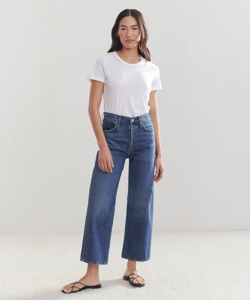A woman stands indoors against a plain light background, wearing a white short-sleeve t-shirt, black sandals, and high-waisted vintage wide-leg denim Ren Jeans.