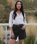 A woman stands outdoors by a white railing and tall grasses, wearing the Stella Short—a pair of high-waisted denim shorts with a belt—and a white long-sleeve buttoned top. Trees are blurred in the background.