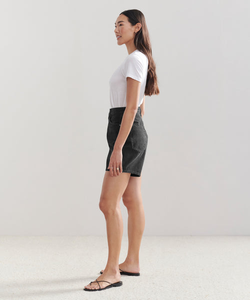 A woman stands in profile against a plain background, wearing Stella Shorts, a white t-shirt, and black sandals. She has long dark hair and a relaxed expression.
