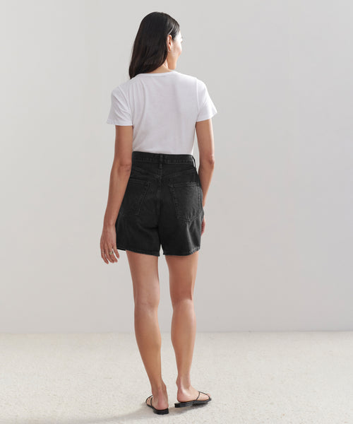 A woman faces away, wearing a white t-shirt, black Stella Short high-waisted denim shorts, and black sandals against a plain light background.